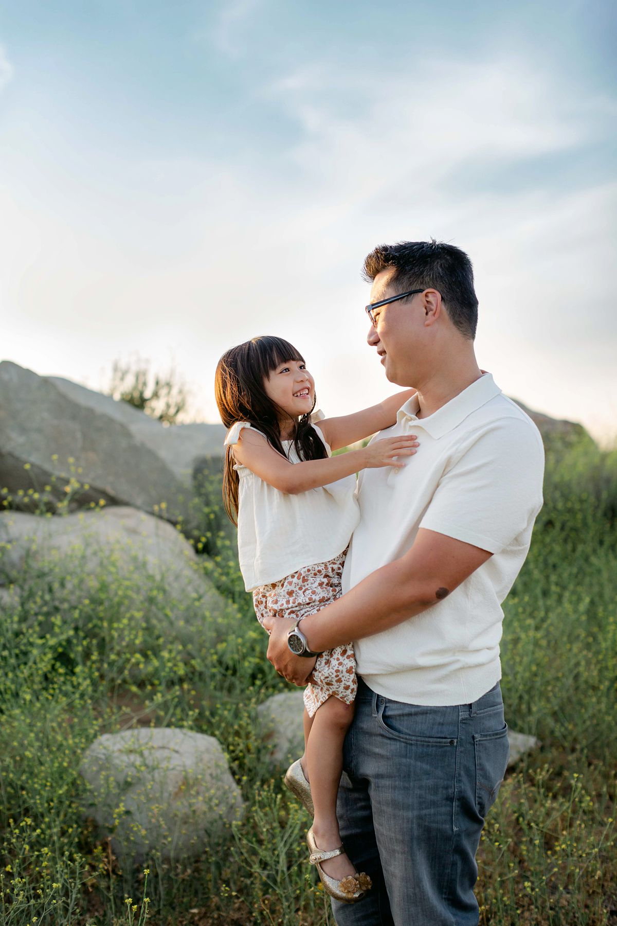 Dad and daughter looking at each other smiling. Boulder and tall grass background.